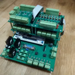 Zestaw trzech zielonych płytek drukowanych ułożonych jedna na drugiej, z licznymi złączami śrubowymi, elementami elektronicznymi i radiatorami, na drewnianym tle.