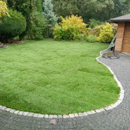 Świeżo ułożony trawnik z rolki, widoczne łączenia, obrzeże z kostki granitowej, w tle różnorodne drzewa i krzewy, obok drewniana altana.