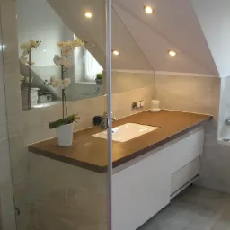 Nowoczesna łazienka na poddaszu z brązowym blatem, białą szafką pod umywalką, kwiatami w doniczce i kabiną prysznicową ze szklanymi drzwiami.