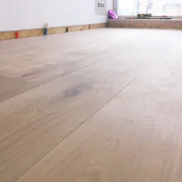 Układanie jasnych paneli podłogowych z widocznym fragmentem okna i narzędziami w tle.