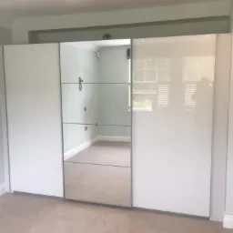 Biała szafa przesuwna z lustrzanym panelem środkowym, odbijająca wnętrze pokoju z jasnymi ścianami i wykładziną, widoczny fragment gniazdka elektrycznego i listwy przypodłogowej.