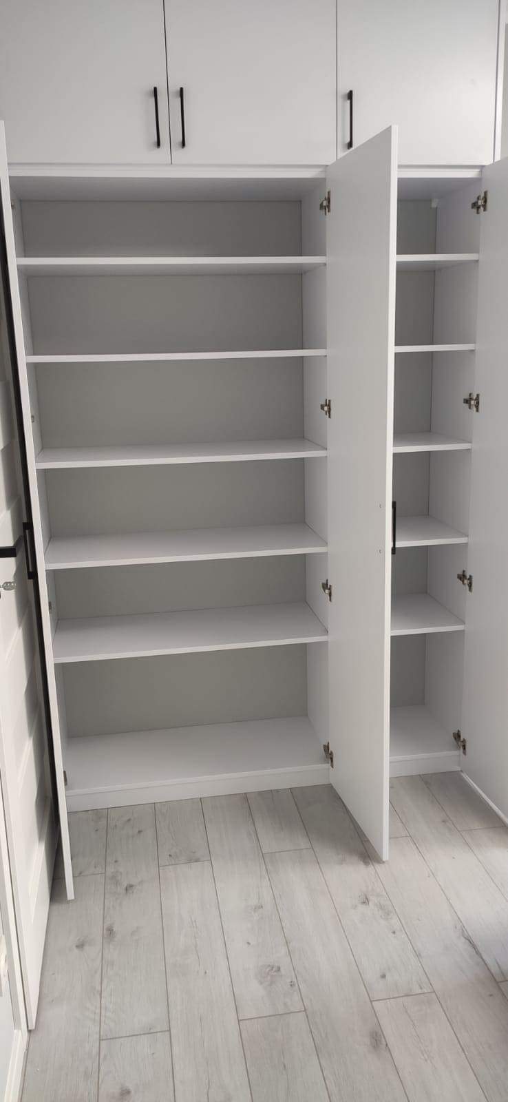 Biała szafa wnękowa z otwartymi drzwiami, widoczne puste półki, czarne uchwyty, podłoga z paneli w odcieniach szarości, realizacja w Police.