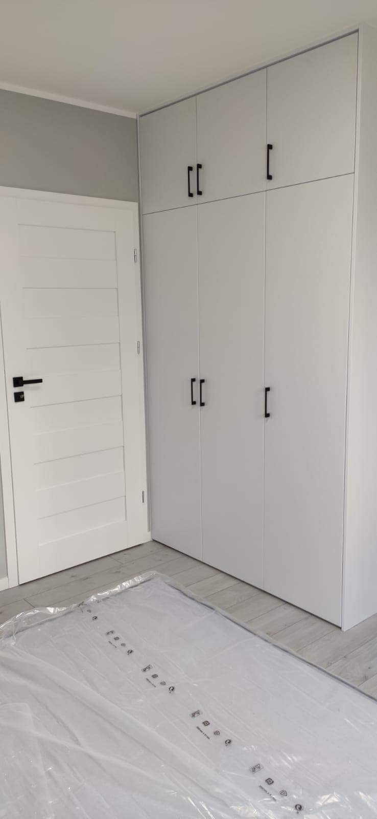 Nowoczesna, biała szafa wnękowa z czarnymi, prostokątnymi uchwytami, obok białe drzwi z poziomymi frezami i czarną klamką, podłoga zabezpieczona folią ochronną.