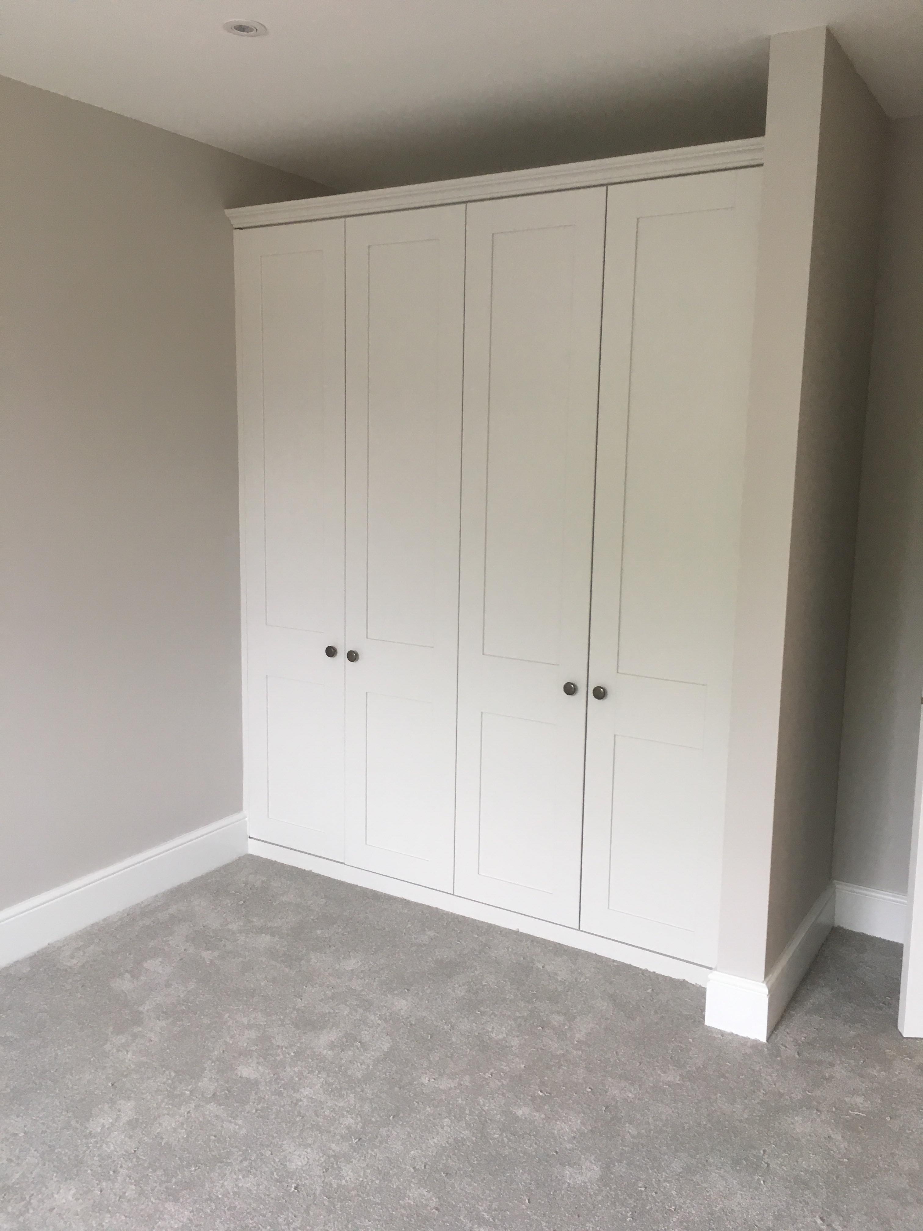 Wbudowana, czterodrzwiowa szafa wnękowa z białymi frontami i okrągłymi, ciemnymi uchwytami, dopasowana do koloru ścian i dywanu w odcieniach szarości.