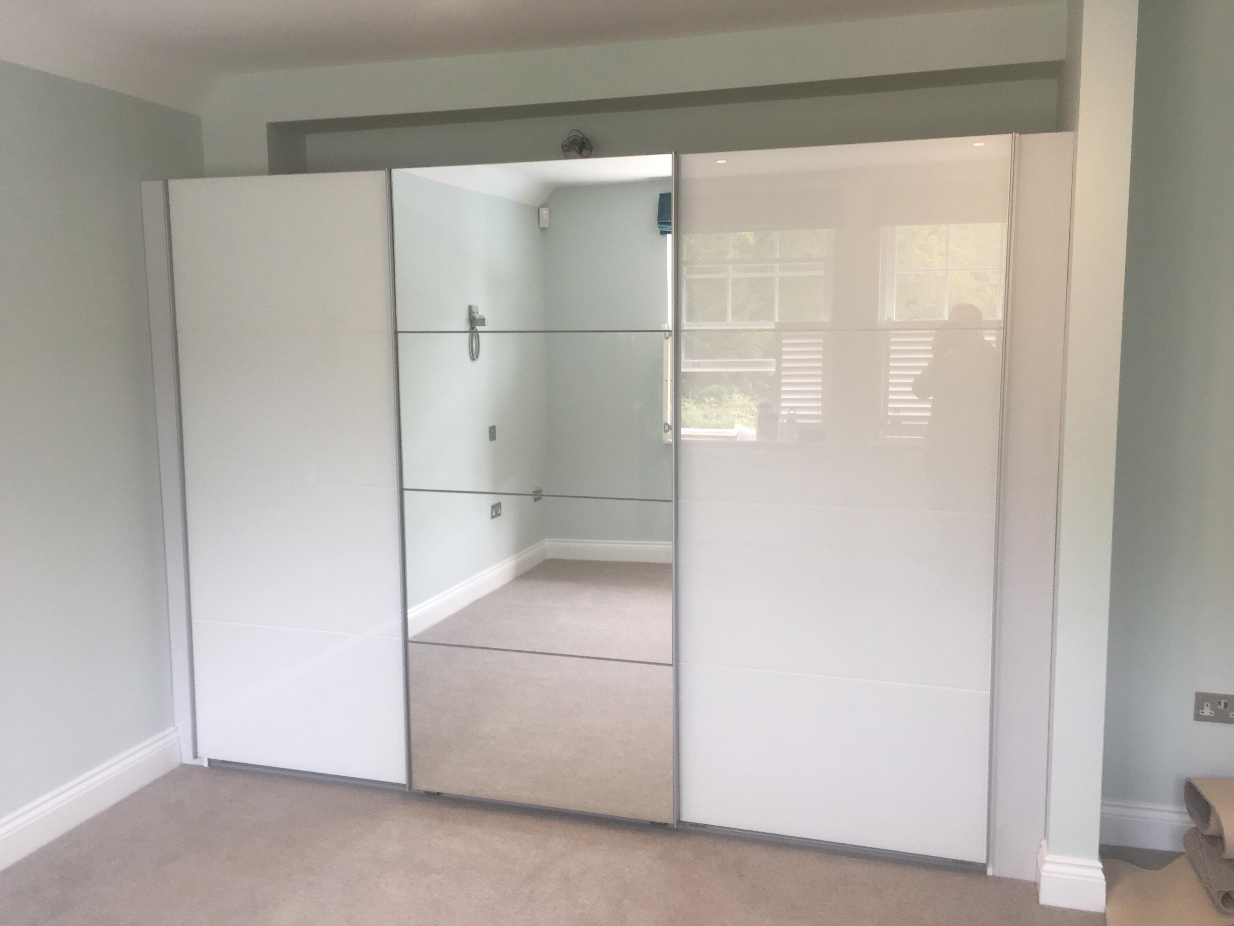 Biała szafa przesuwna z lustrzanym panelem środkowym, odbijająca wnętrze pokoju z jasnymi ścianami i wykładziną, widoczny fragment gniazdka elektrycznego i listwy przypodłogowej.