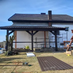 Budynek z dobudowanym drewnianym zadaszeniem w trakcie prac, widoczne rusztowania, ocieplenie styropianem, dach z dachówki ceramicznej, narzędzia i materiały budowlane na trawniku, w tle ogrodzenie.