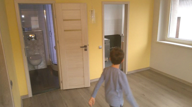 Żółta ściana w pokoju z dwiema otwartymi, drewnianymi drzwiami do łazienki i kuchni, widoczny chłopiec w tle.