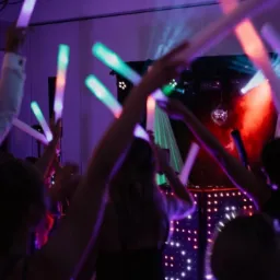 Roztańczony tłum z podniesionymi świetlnymi pałeczkami na imprezie, w tle scena z dyskotekowymi kulami i oświetleniem punktowym.