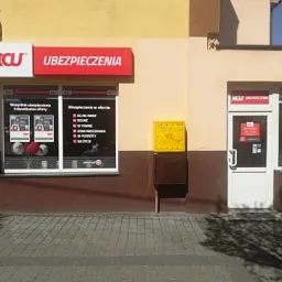 Fasada budynku z szyldem firmy ubezpieczeniowej RCW z ofertą różnorodnych polis, w tym OC, rolne, domów, na życie. Żółta skrzynka gazowa obok wejścia.