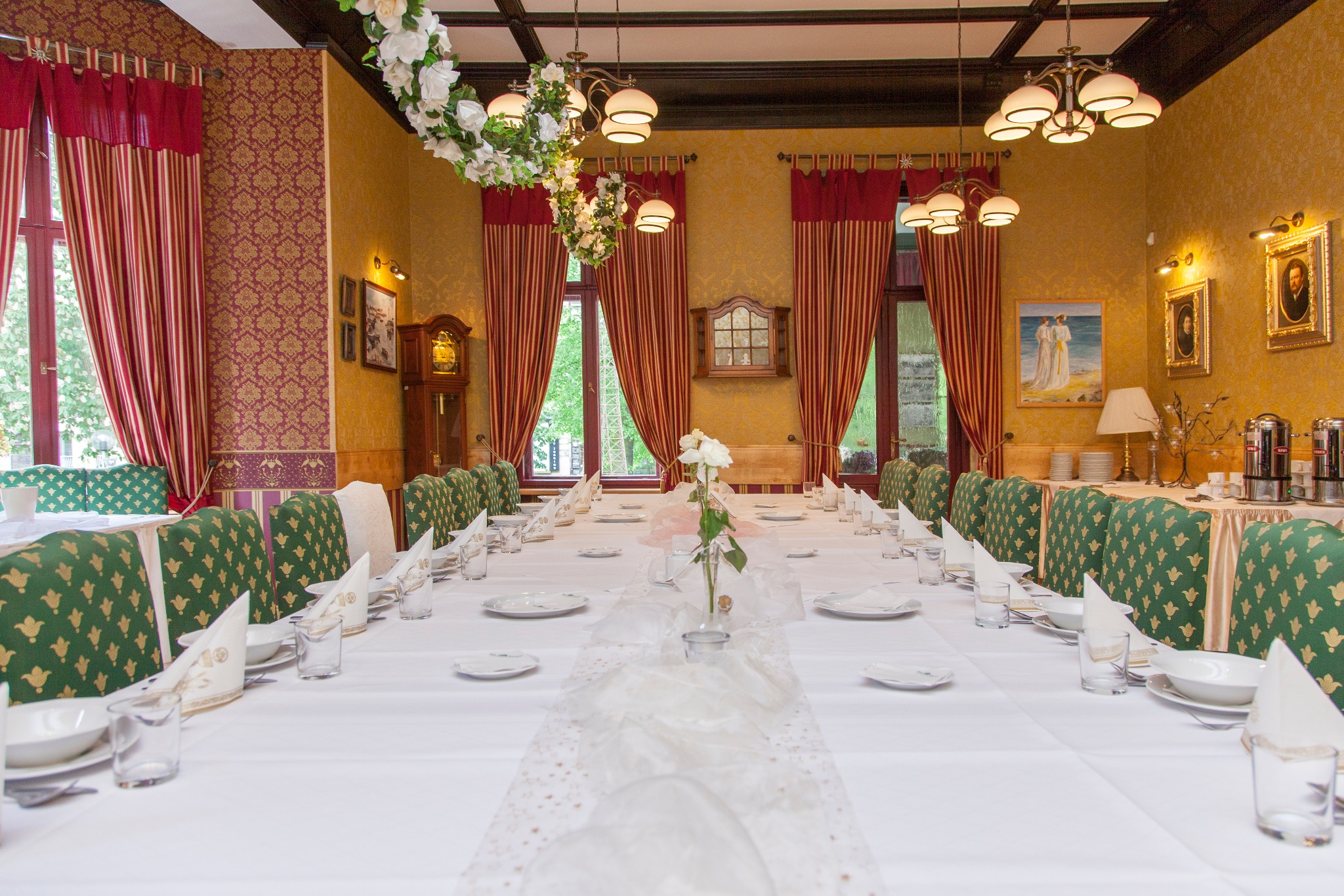 Długi stół nakryty białym obrusem, zastawiony talerzami, szklankami i serwetkami, udekorowany kwiatami, otoczony zielonymi krzesłami w sali z wzorzystymi tapetami i dekoracyjnymi lampami.