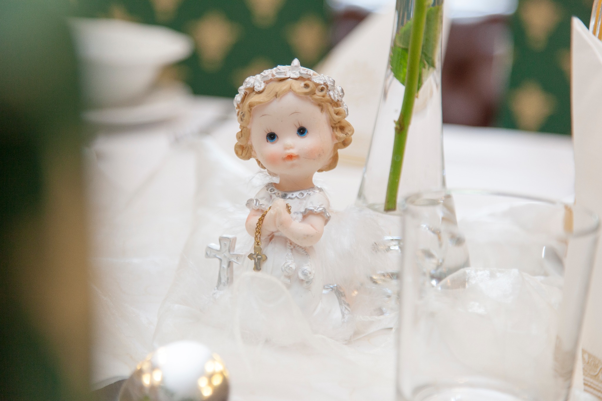Dekoracja stołu komunijnego: figurka dziewczynki w białej sukience z krzyżykiem w dłoniach, otoczona białymi piórkami, z rozetą na głowie, obok szklanka i wazon z różą.