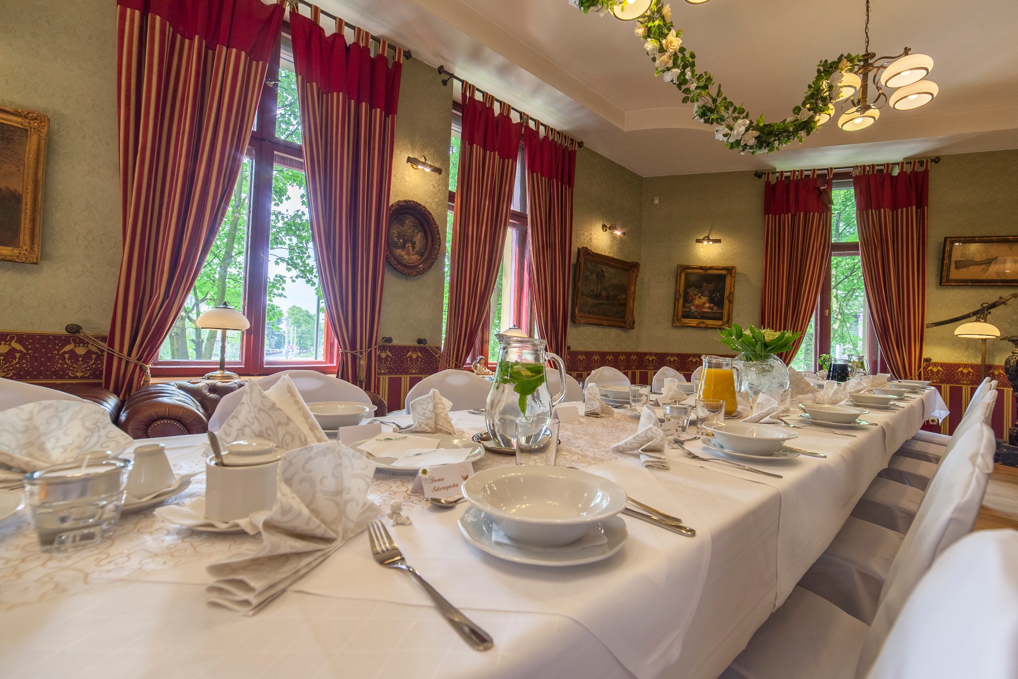 Elegancko nakryty stół z białą zastawą, dzbankiem z wodą i miętą, dekoracjami kwiatowymi, w tle okna z czerwono-białymi zasłonami i obrazy w ozdobnych ramach.