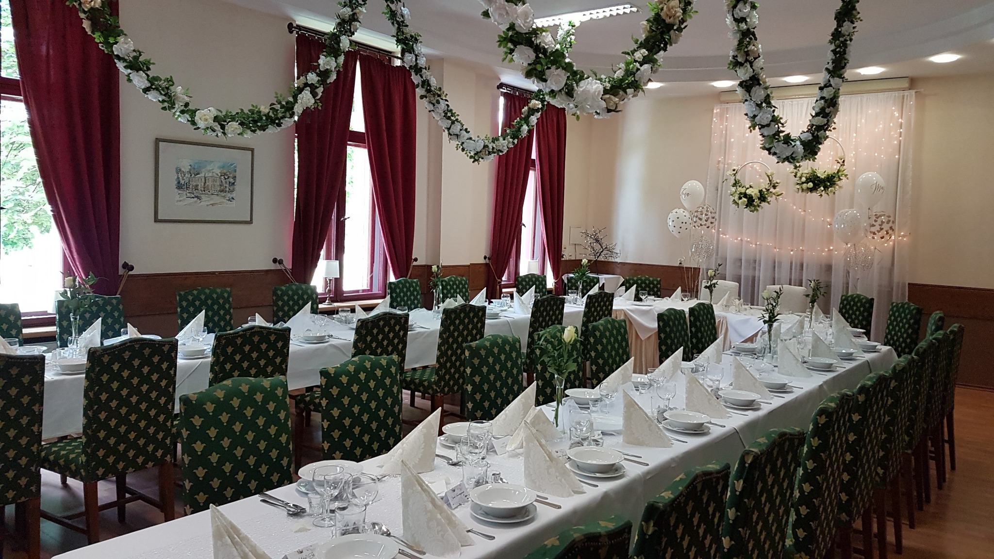 Uroczysta sala z długim stołem nakrytym białym obrusem, zastawionym porcelaną i szkłem, udekorowana kwiatowymi girlandami i balonami, z krzesłami o zielonej tapicerce w złote wzory.