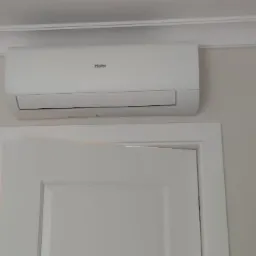 Biały klimatyzator Haier zamontowany nad drzwiami w pokoju z dekoracyjnymi listwami ściennymi i lampą ścienną w stylu art deco. Minimalistyczny design.