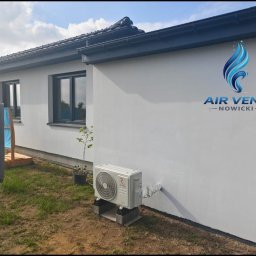 AIR VENT Nowicki Rekuperacje Sp. z o.o. - Biała jednostka zewnętrzna systemu chłodzenia/grzania marki Revent, zamontowana na betonowych bloczkach przy białej elewacji nowoczesnego domu z ciemnymi oknami i dachem. Widoczny trawnik i małe...