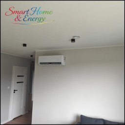 AIR VENT Nowicki Rekuperacje Sp. z o.o. - Nowoczesny biały klimatyzator ścienny zamontowany na beżowej ścianie w jasnym wnętrzu, z czarnymi lampami punktowymi i białymi drzwiami w tle.