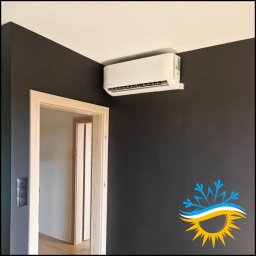 AIR VENT Nowicki Rekuperacje Sp. z o.o. - Biała jednostka klimatyzacji ściennej zamontowana w narożniku pokoju z ciemnoszarymi ścianami i białym sufitem, obok jasna drewniana ościeżnica drzwiowa.