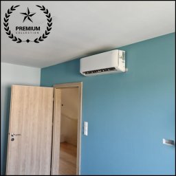 AIR VENT Nowicki Rekuperacje Sp. z o.o. - Biały klimatyzator ścienny zamontowany na turkusowej ścianie, obok otwartych jasnych drewnianych drzwi. W tle widoczne wnętrze pomieszczenia.