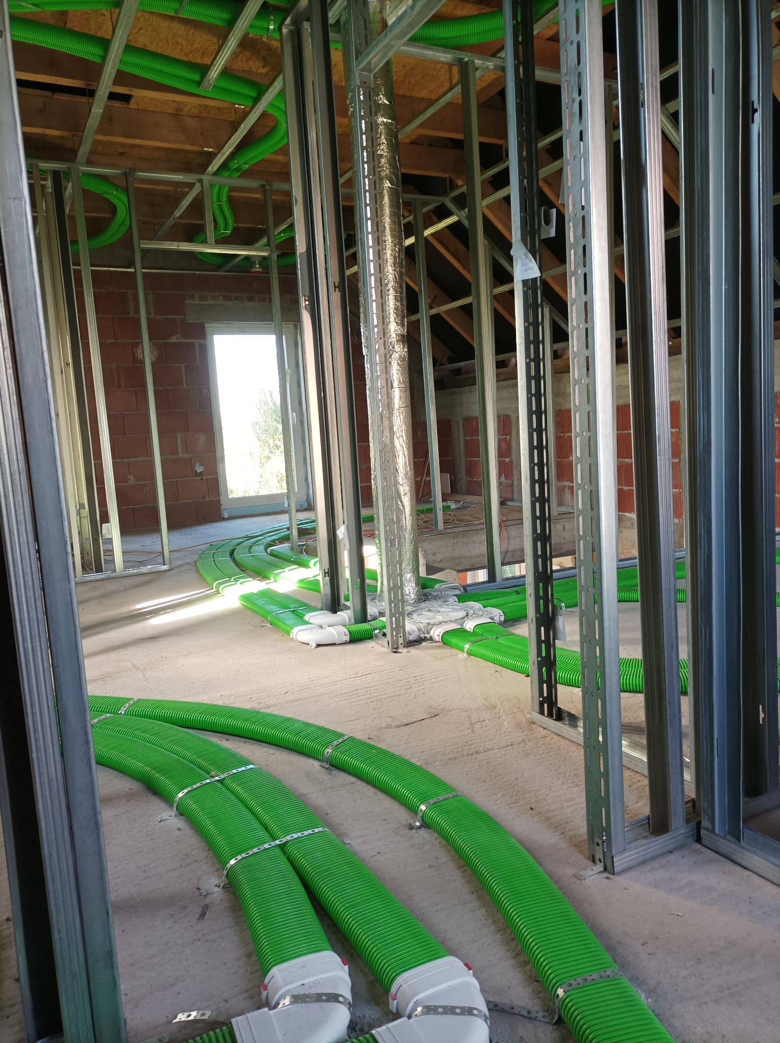 Wnętrze w trakcie montażu rekuperacji: widoczne zielone rury wentylacyjne na podłodze i pod sufitem, metalowa konstrukcja ścian działowych, surowa cegła i drewniana więźba dachowa.