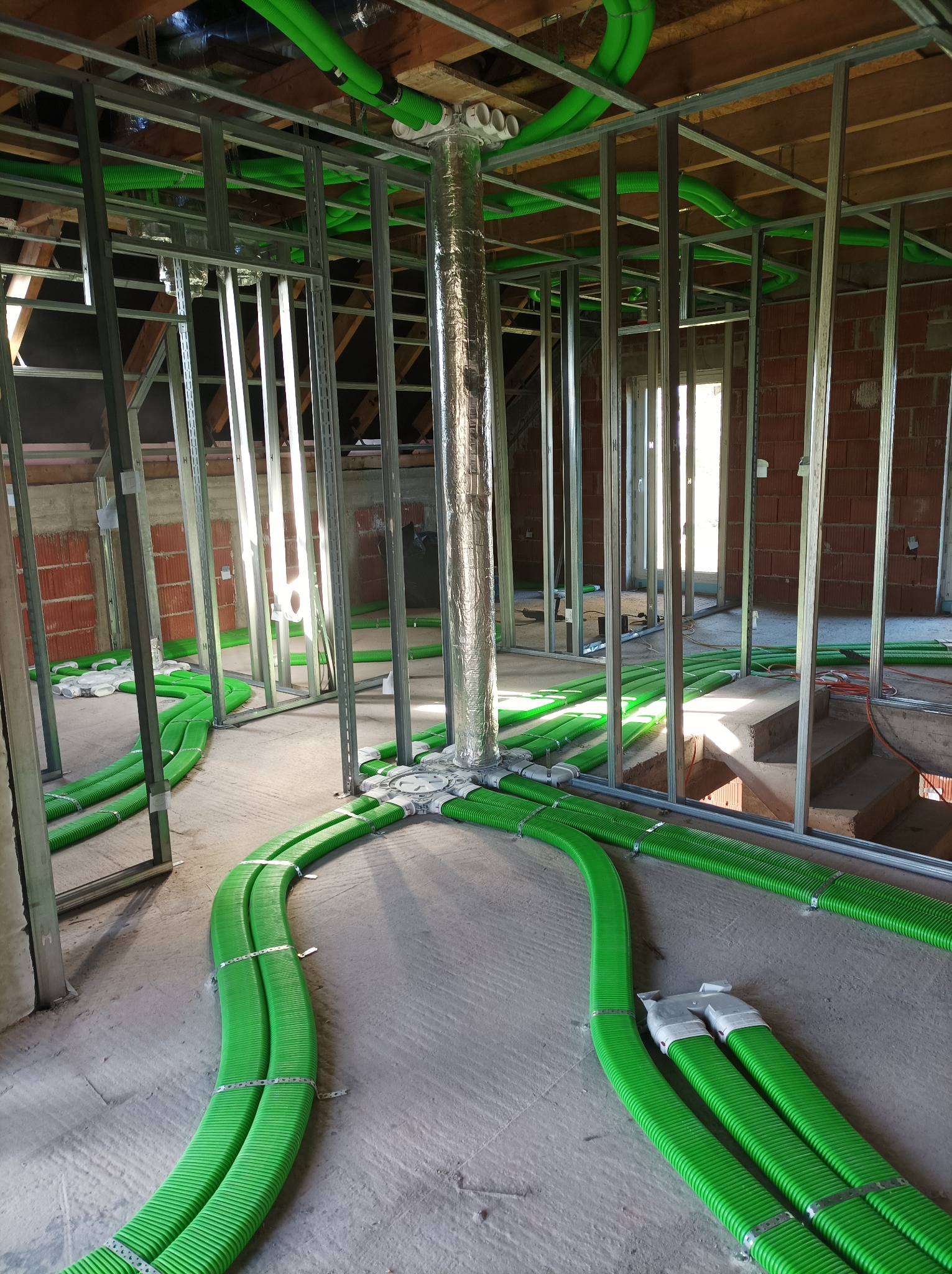 Wnętrze w trakcie budowy z widoczną instalacją wentylacyjną z zielonych, karbowanych rur prowadzonych po podłodze i pod stropem, metalowa konstrukcja ścian działowych w tle, rury łączą się...