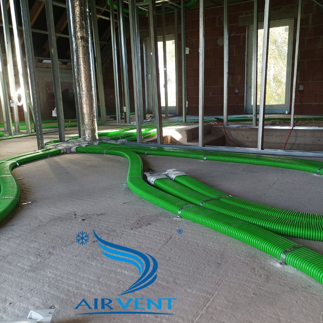 Wnętrze w trakcie montażu wentylacji: zielone, karbowane rury wentylacyjne prowadzone po podłodze, stalowa konstrukcja ścian działowych, okno w tle, widoczne logo firmy AIRVENT.