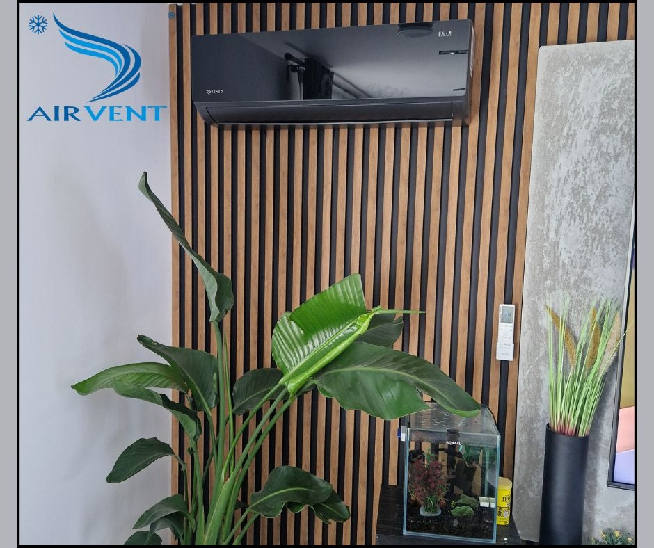 Czarna klimatyzacja ścienna z pilotem, zamontowana na dekoracyjnej ścianie z drewnianych listew, z rośliną doniczkową i akwarium w pomieszczeniu.