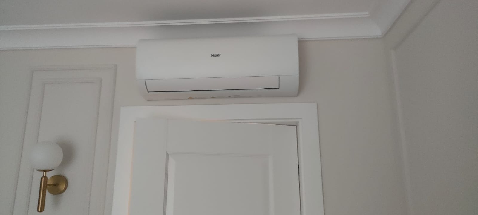 Biały klimatyzator Haier zamontowany nad drzwiami w pokoju z dekoracyjnymi listwami ściennymi i lampą ścienną w stylu art deco. Minimalistyczny design.