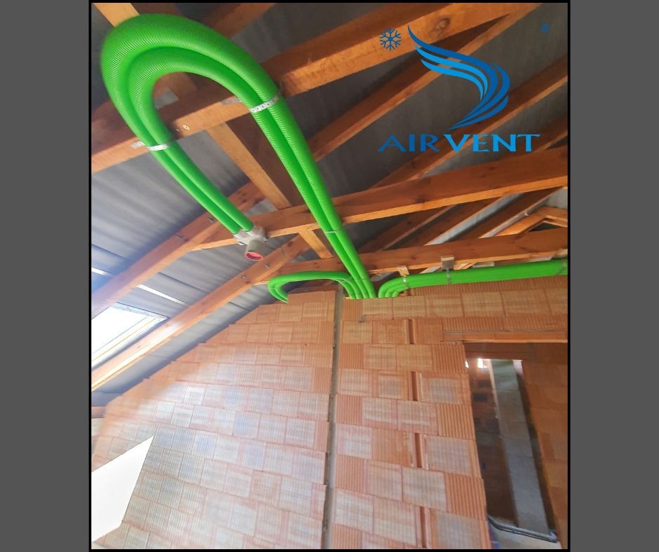 Zielone rury wentylacyjne pod stropem z drewnianych belek, widoczne ceglane ściany w surowym stanie i logo firmy AirVent na tle dachu.