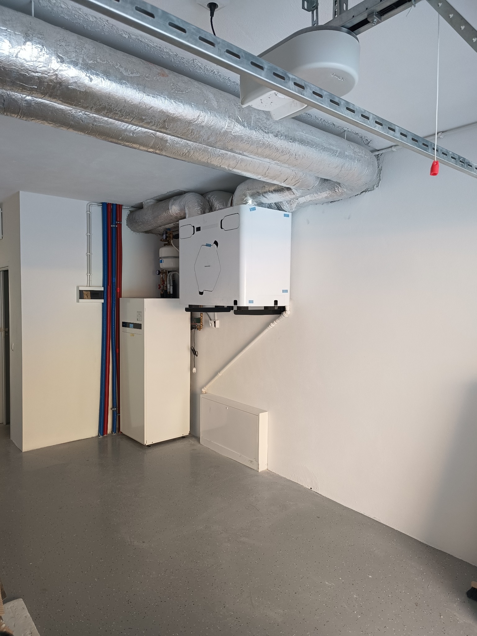 Biała jednostka rekuperacyjna zamontowana na ścianie, z widocznymi srebrnymi rurami wentylacyjnymi pod sufitem. Instalacja w pomieszczeniu z szarą podłogą.