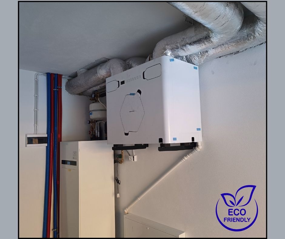 Biała jednostka rekuperacyjna zamontowana na ścianie, z widocznymi rurami wentylacyjnymi w izolacji. Na ścianie widoczne rury instalacyjne w kolorach niebieskim i czerwonym.