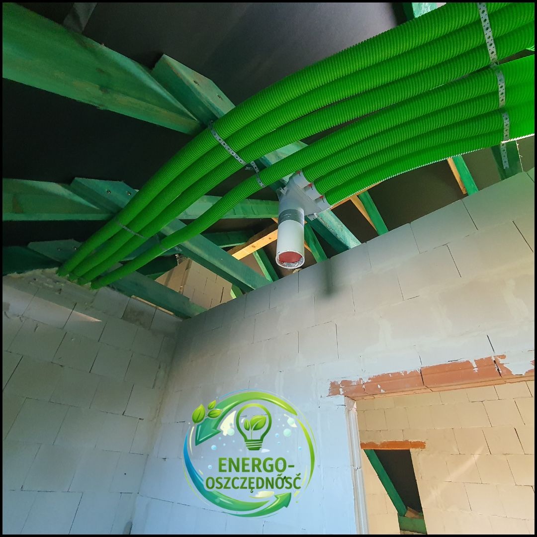 Zielone rury wentylacyjne pod sufitem poddasza, zamocowane do drewnianej konstrukcji. Widoczny biały rozdzielacz. Ściany z białego bloczka. Logo firmy w rogu.