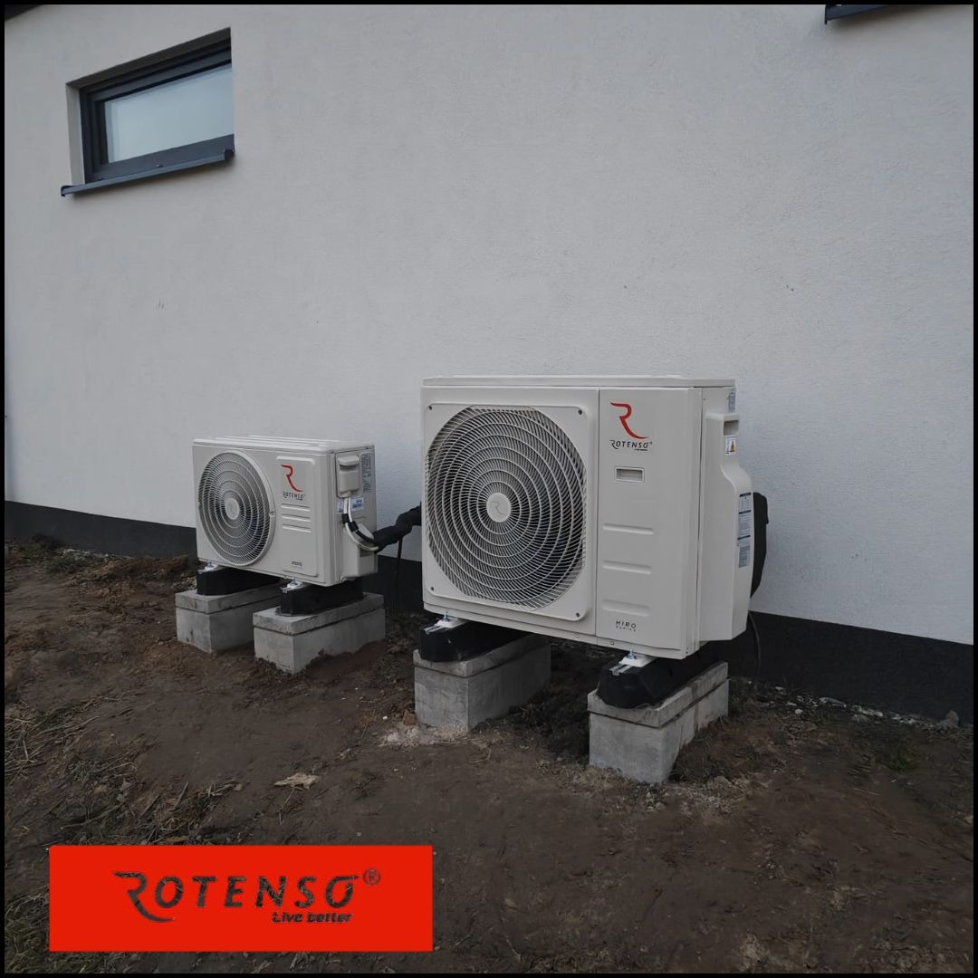 Dwie jednostki klimatyzacji Rotenso HIRO na betonowych podstawach przy białej ścianie budynku z małym oknem. Widok z zewnątrz, instalacja przy ziemi.