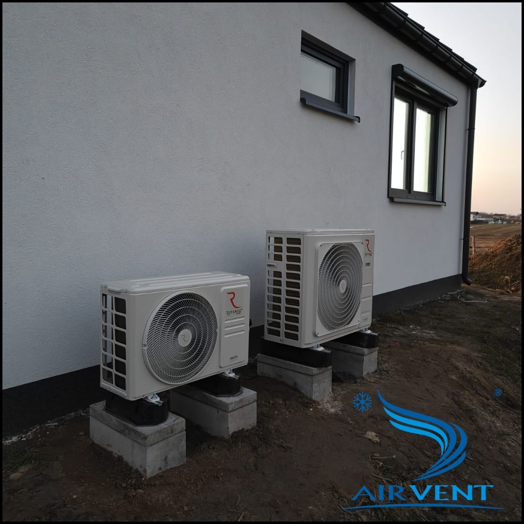 Dwa białe klimatyzatory Rotenso na betonowych podstawach przy ścianie domu z oknami. Widoczne logo Air Vent w rogu. Ujęcie z dołu, ciepłe światło zachodzącego słońca.