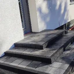 Schody wejściowe do domu wyłożone szarą kostką brukową, widoczne niezakończone prace ziemne wokół.