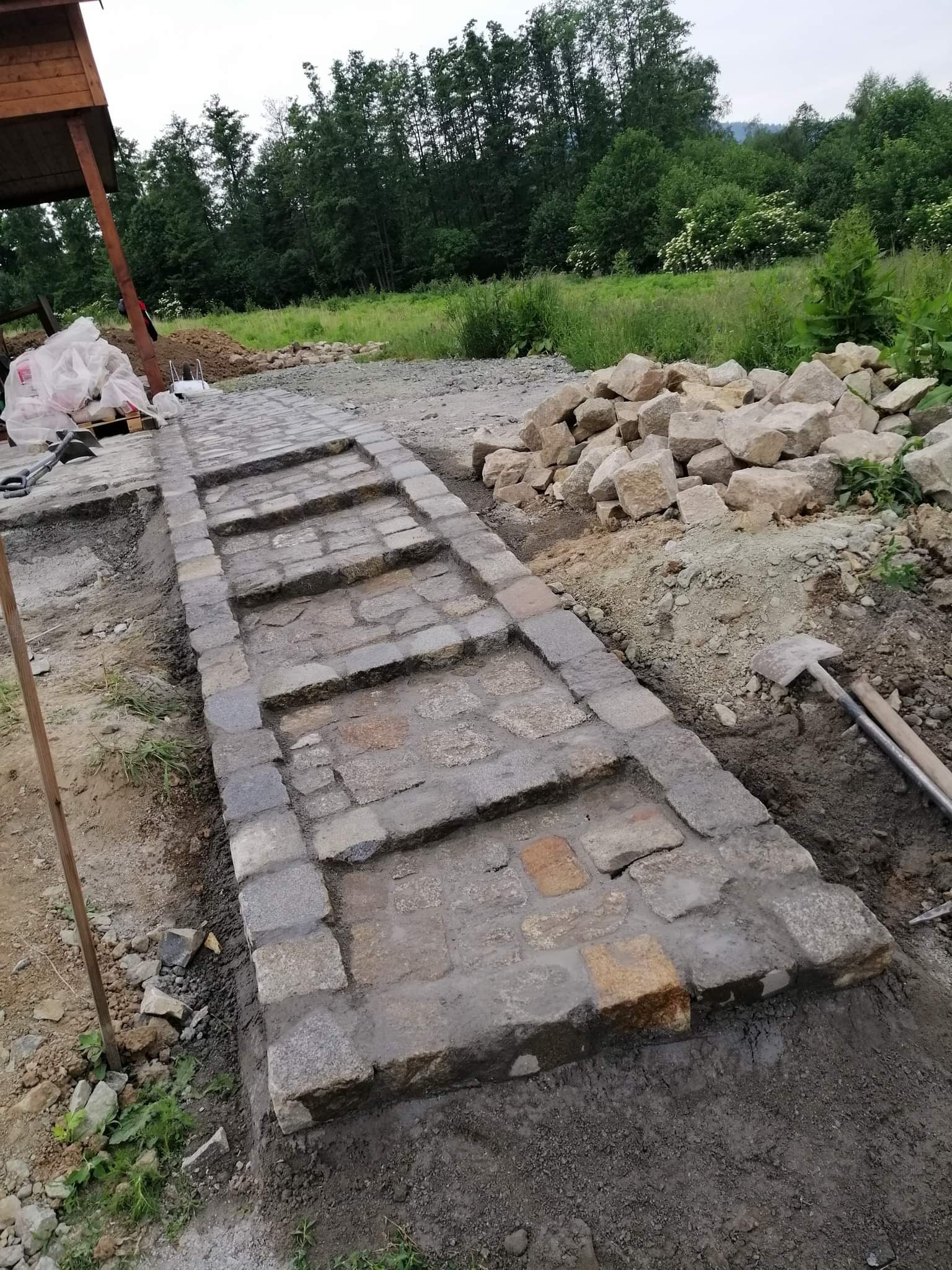 Układanie kamiennej ścieżki schodkowej w ogrodzie, widoczne narzędzia i materiały budowlane, tło z zielenią.