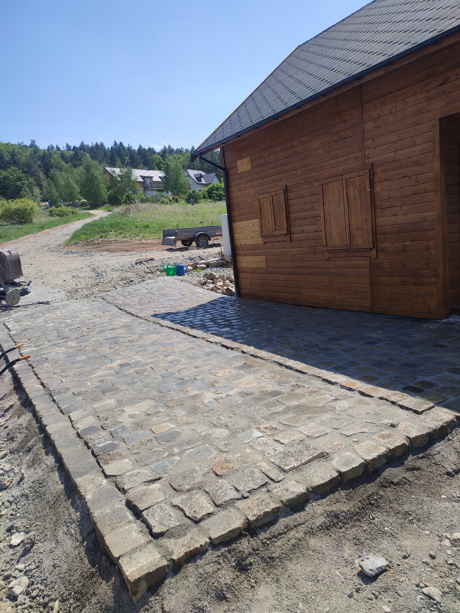 Świeżo ułożona kostka brukowa z granitu w odcieniach szarości i beżu, prowadząca do drewnianego domku z brązowymi okiennicami, w tle zielone wzgórza i błękitne niebo.