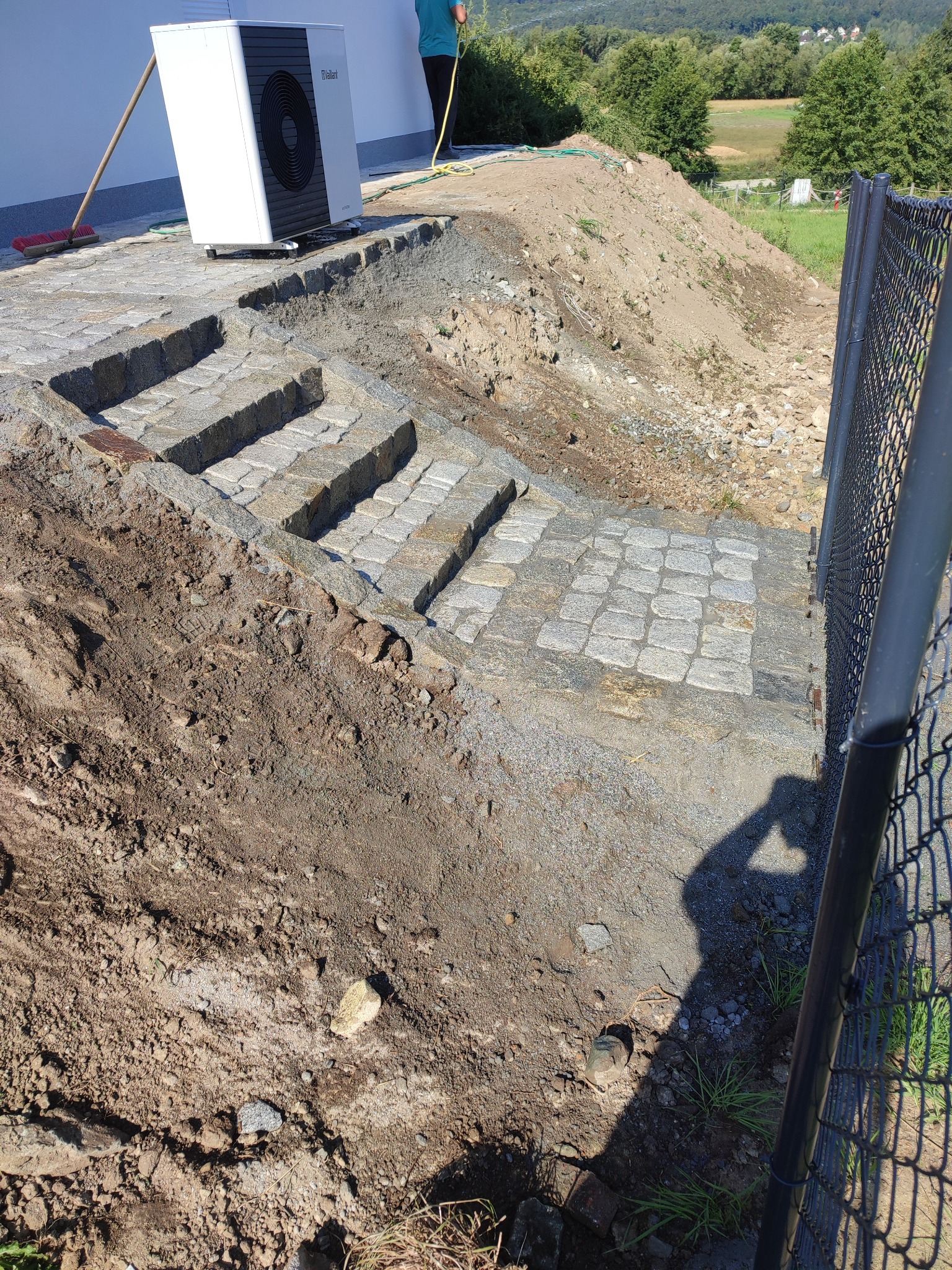 Schody zewnętrzne z kostki granitowej w trakcie budowy, obok zamontowana pompa ciepła, widoczny fragment ogrodzenia z siatki i sterta ziemi.