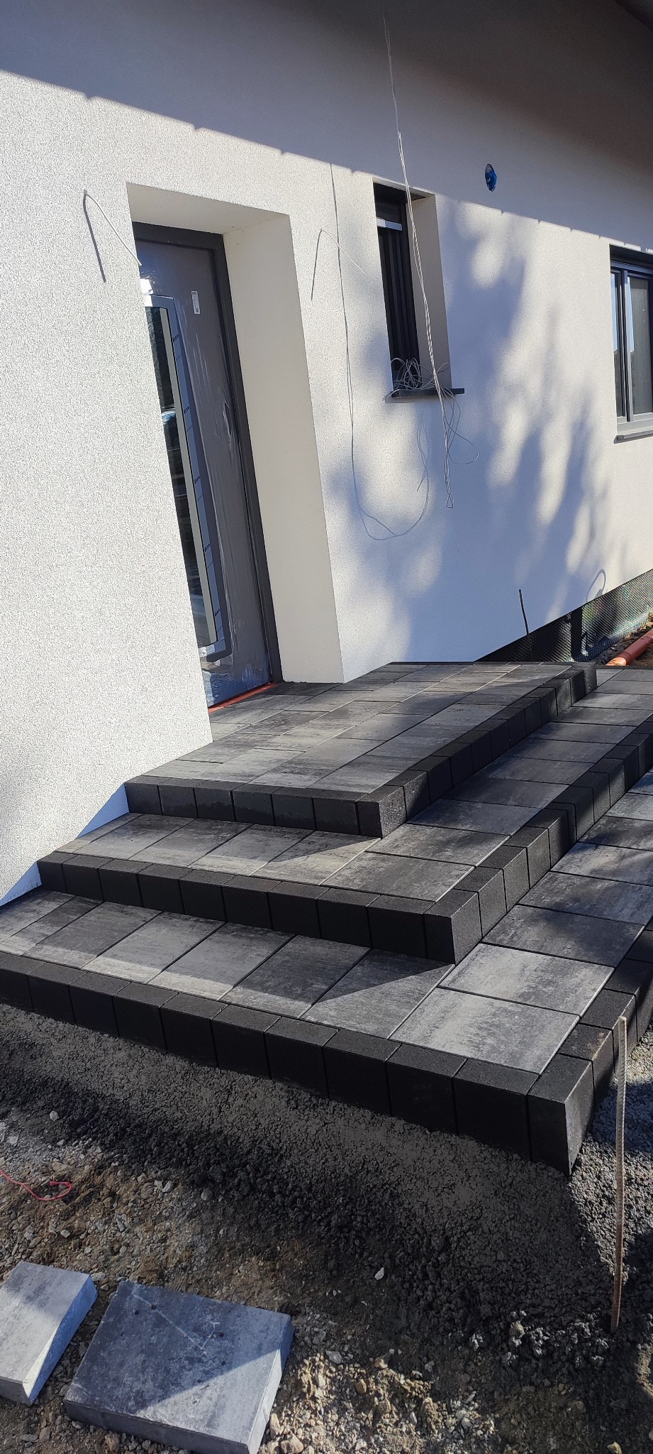 Schody wejściowe do domu wyłożone szarą kostką brukową, widoczne niezakończone prace ziemne wokół.