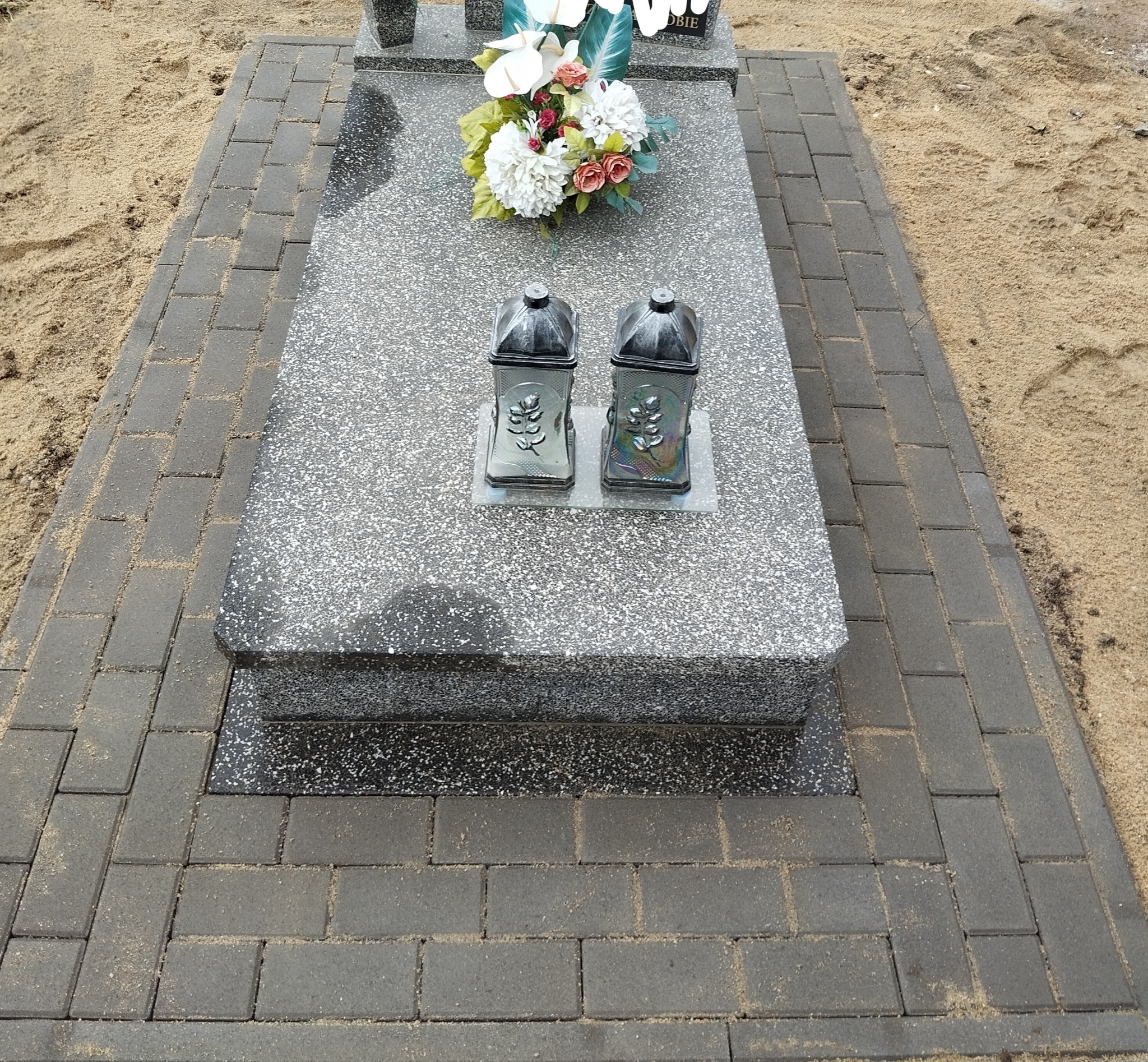 Granitowy nagrobek otoczony szarą kostką brukową, z dwoma lampionami i bukietem sztucznych kwiatów na płycie.