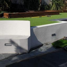 Onyx - Nowoczesny ogród z tarasem wyłożonym szarym kamieniem, białymi ścianami z oświetleniem punktowym i sztuczną trawą na różnych poziomach, fragment rolki sztucznej trawy na posadzce.