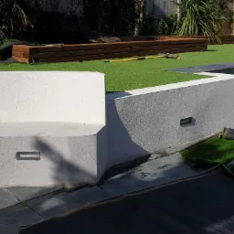 Nowoczesny ogród z tarasem wyłożonym szarym kamieniem, białymi ścianami z oświetleniem punktowym i sztuczną trawą na różnych poziomach, fragment rolki sztucznej trawy na posadzce.