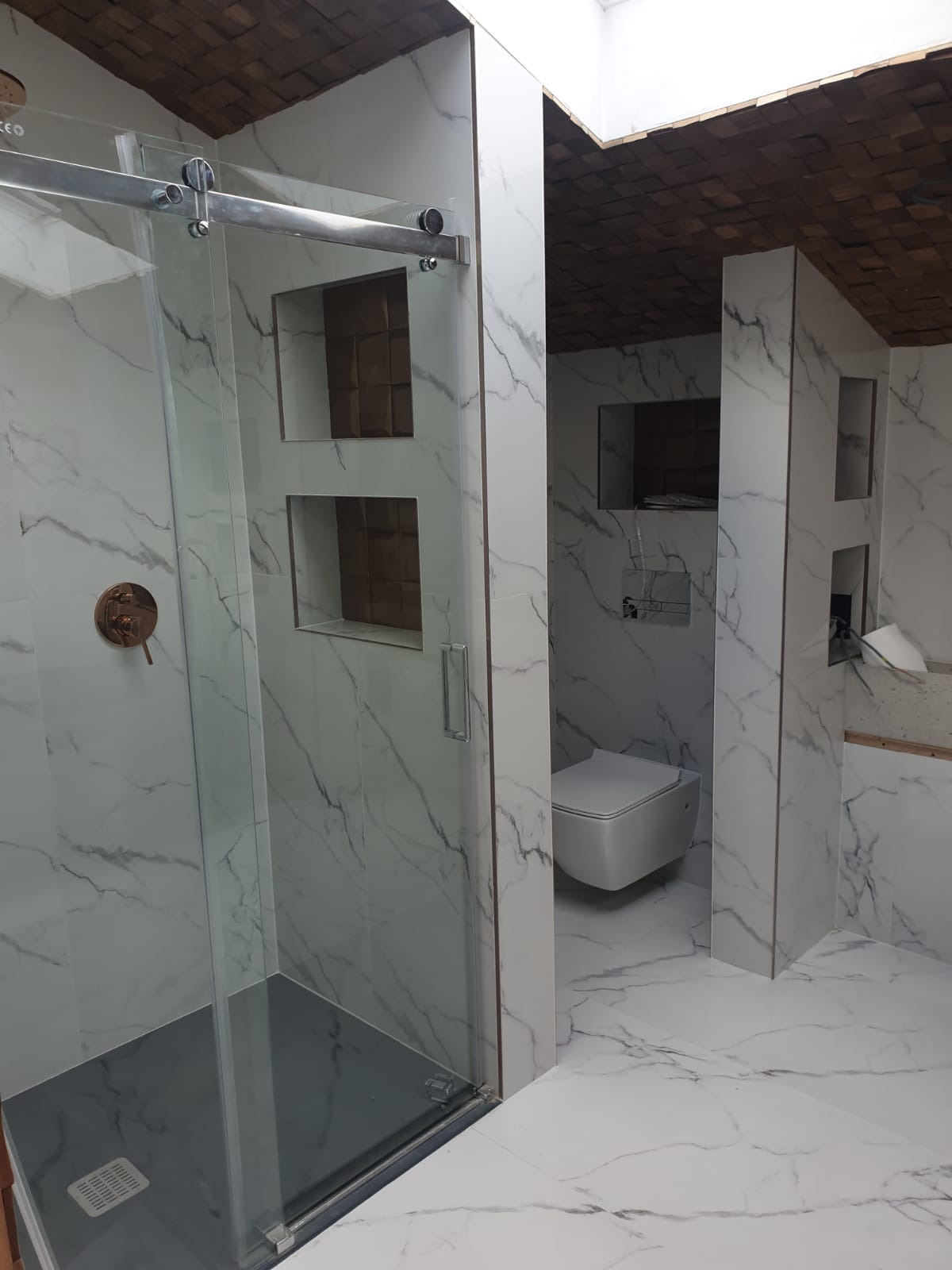 Wykończona łazienka z kabiną prysznicową ze szklanymi drzwiami przesuwnymi, marmurowe płytki na ścianach i podłodze, drewniany sufit z oknem dachowym, wisząca toaleta i wnęki w ścianie.