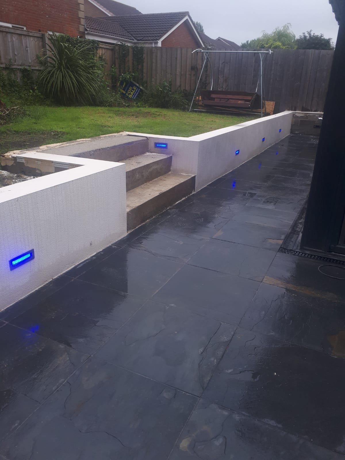 Nowoczesny taras z ciemnej, błyszczącej kostki, oświetlony niebieskimi lampami w ścianie, z widocznymi schodami i fragmentem ogrodu z huśtawką.