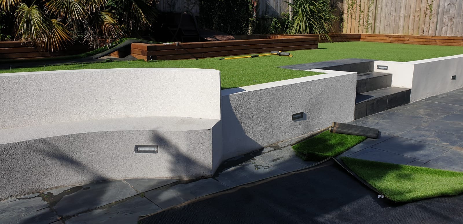 Nowoczesny ogród z tarasem wyłożonym szarym kamieniem, białymi ścianami z oświetleniem punktowym i sztuczną trawą na różnych poziomach, fragment rolki sztucznej trawy na posadzce.