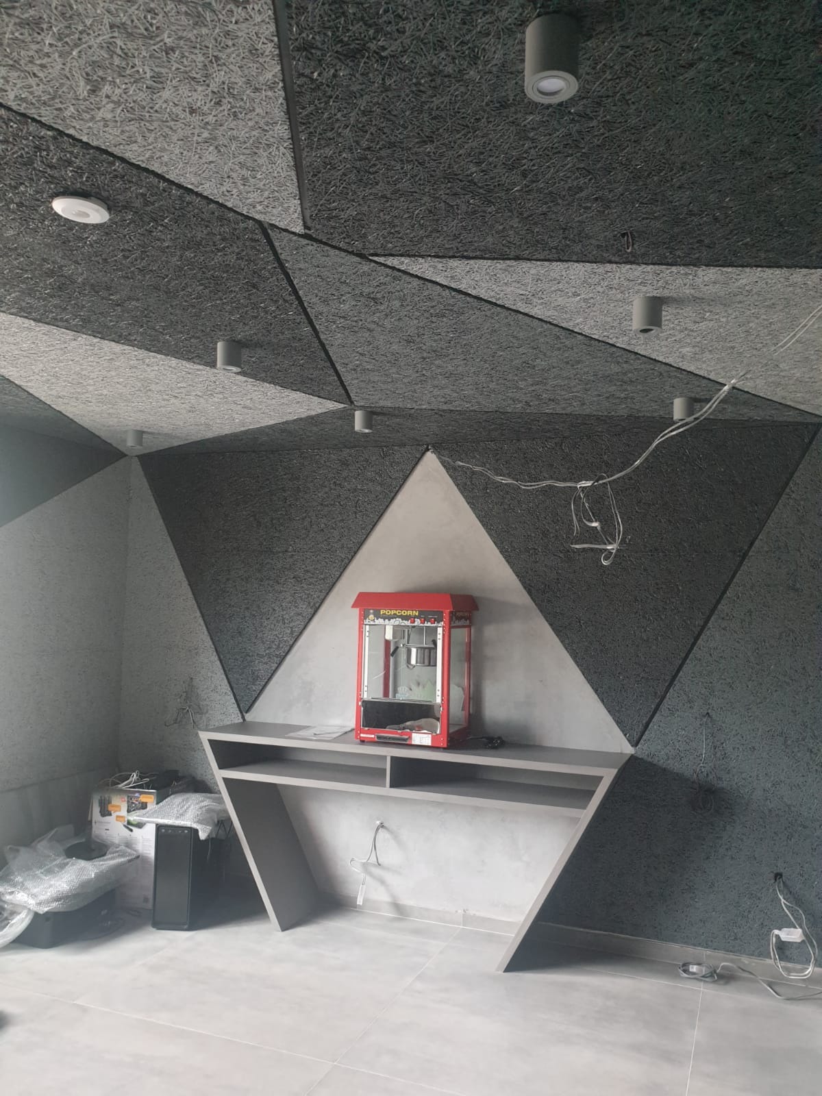 Pokój z akustyczną okładziną ścian i sufitu w geometryczne wzory, szaro-biała kolorystyka, oświetlenie punktowe, czerwony automat do popcornu na półce, widoczne kable i sprzęt elektroniczny.