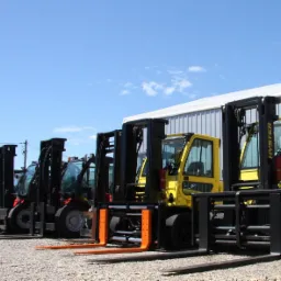 Rząd wózków widłowych różnych typów i kolorów, w tym żółty Hyster, zaparkowanych na żwirowym placu w Andrychowie, z budynkiem magazynowym w tle.