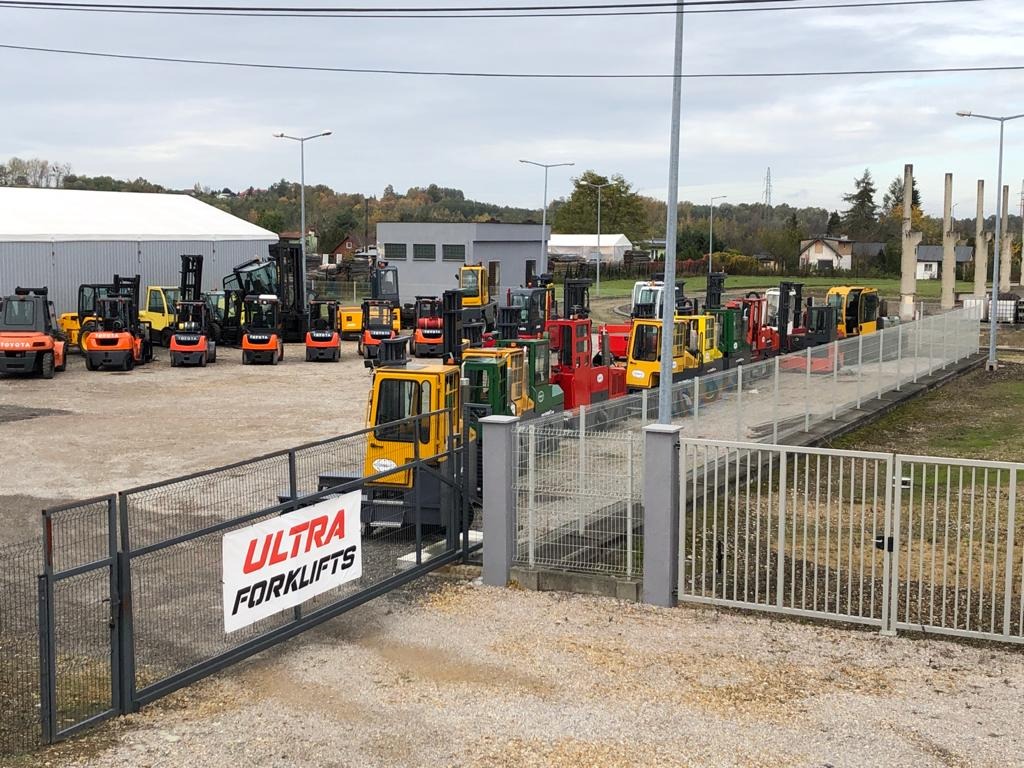 Szeroki kadr na plac z różnorodnymi wózkami widłowymi i ładowarkami teleskopowymi, widok zza ogrodzenia z banerem 'ULTRA FORKLIFTS', pochmurne niebo w tle.