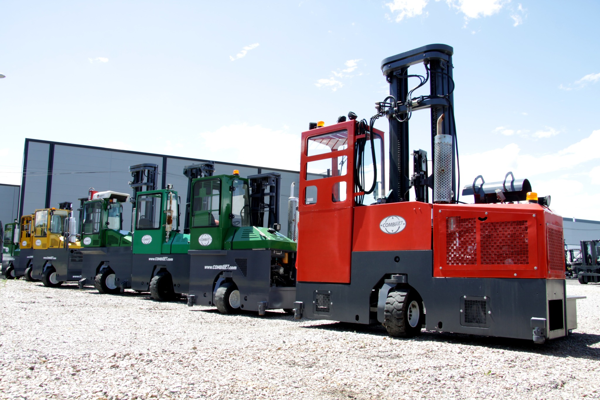 Rząd nowych wózków widłowych wielokierunkowych marki Combilift w różnych kolorach (czerwony, zielony, żółty) na żwirowym placu, z budynkiem magazynowym w tle.