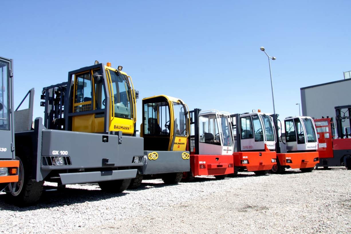 Rząd nowych, różnokolorowych wózków widłowych marki Baumann zaparkowanych na żwirowym placu pod błękitnym niebem.