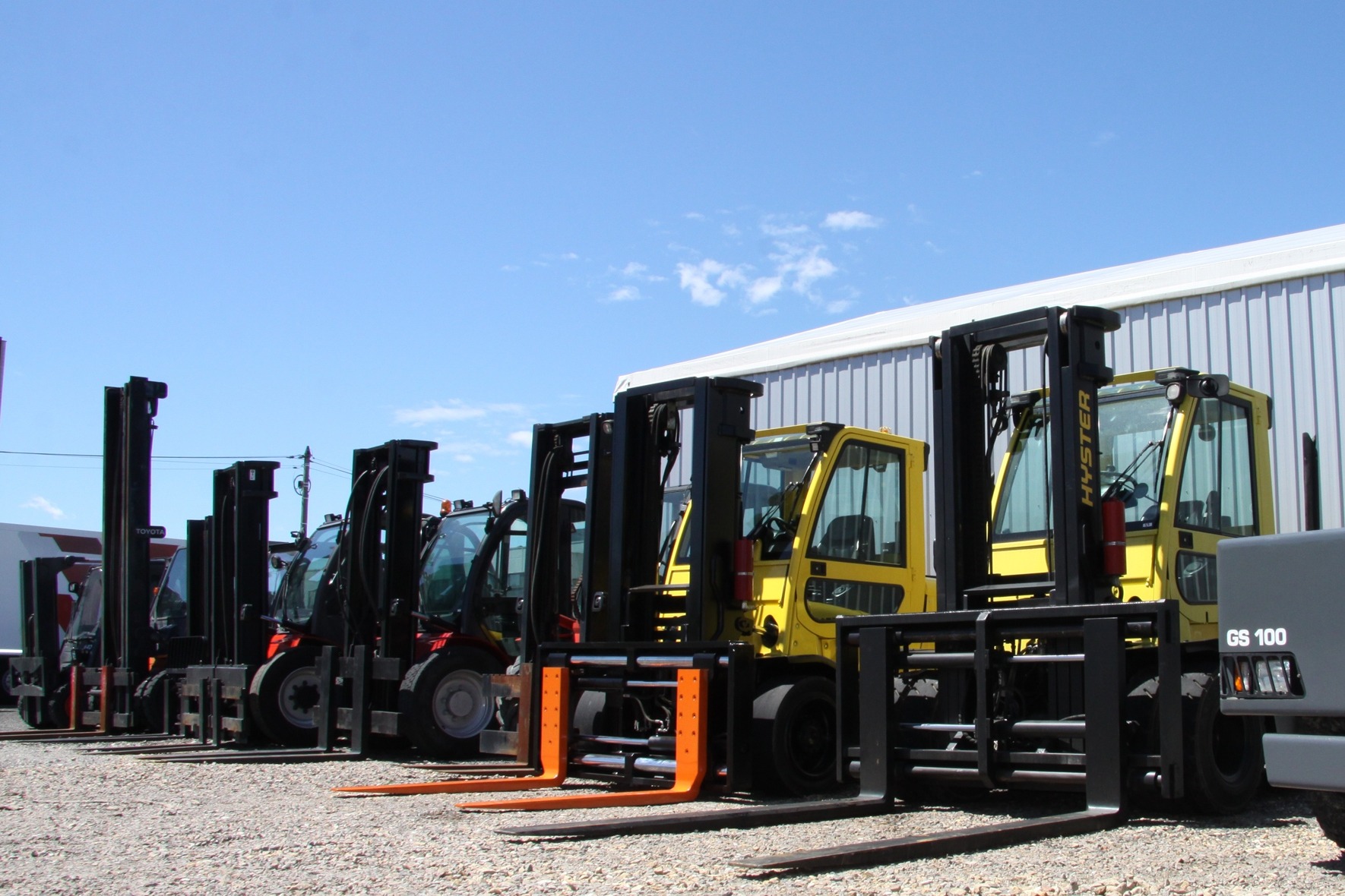 Rząd wózków widłowych różnych typów i kolorów, w tym żółty Hyster, zaparkowanych na żwirowym placu w Andrychowie, z budynkiem magazynowym w tle.